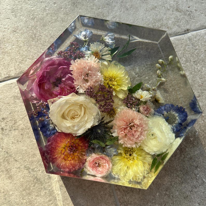 May include: A hexagonal resin paperweight with a variety of pressed flowers in shades of pink, white, yellow, and blue.