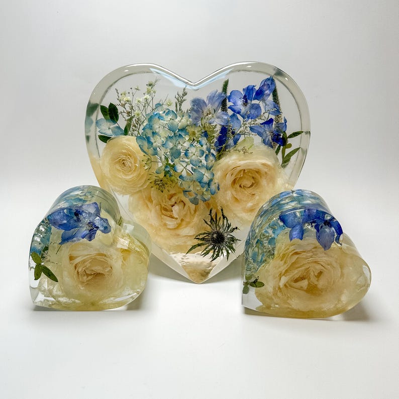 May include: Three heart-shaped resin ornaments with preserved flowers. The largest ornament is a heart with a bouquet of white roses and blue flowers. The two smaller ornaments are hearts with a single white rose and blue flowers.