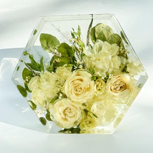 May include: A hexagonal clear resin block with a bouquet of preserved white roses and greenery inside.
