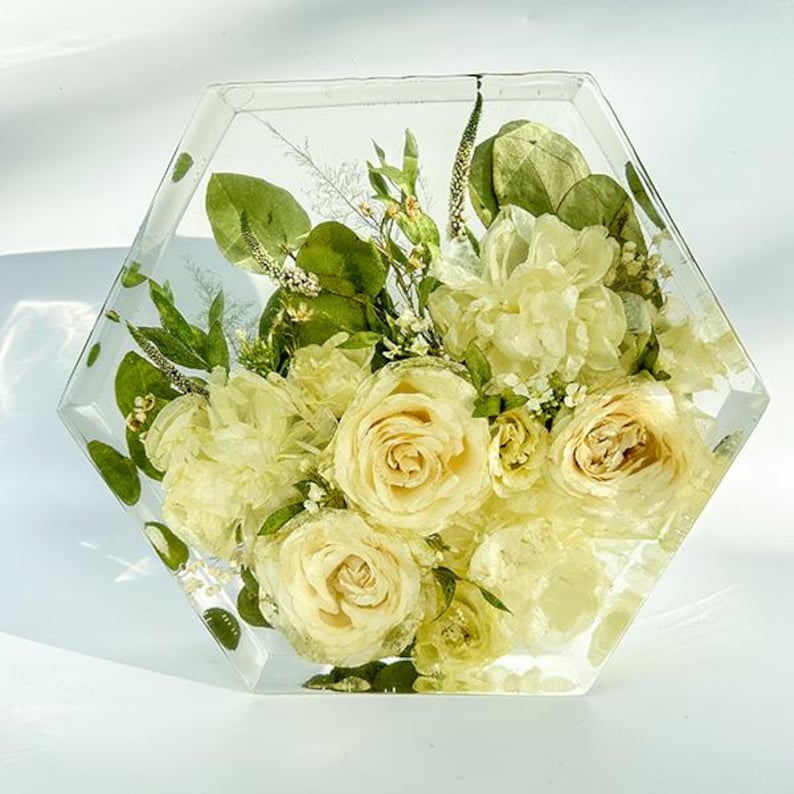 May include: A hexagonal clear resin block with a bouquet of preserved white roses and greenery inside.