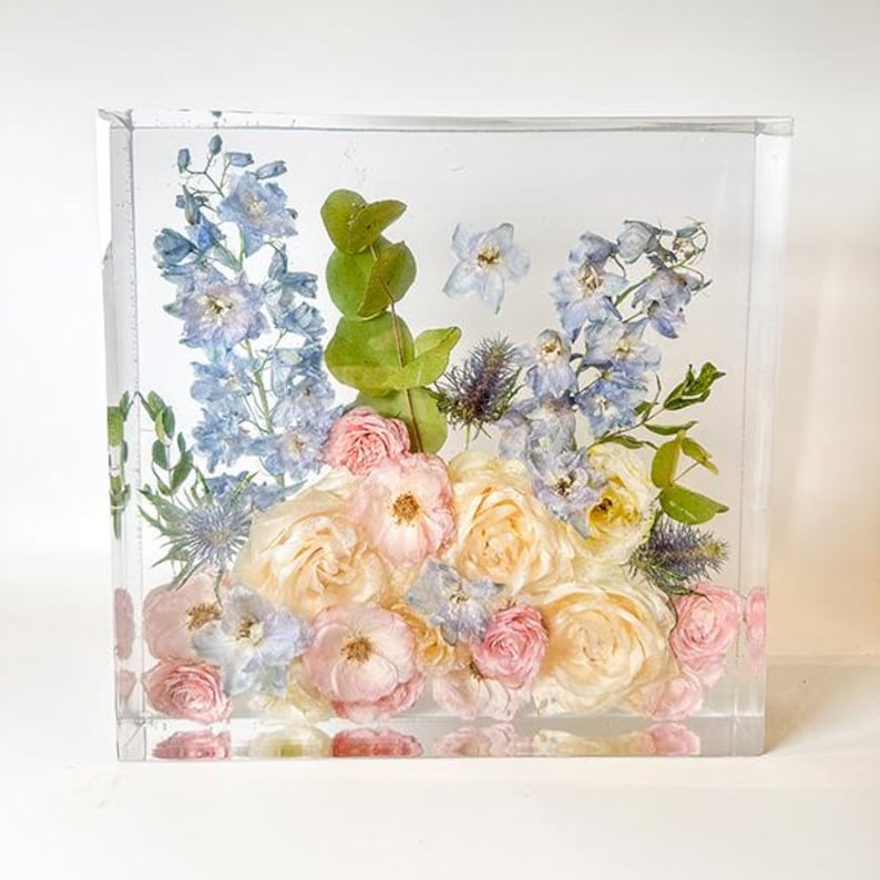 Puede incluir: Un cubo transparente lleno de flores preservadas. Las flores son una mezcla de azul claro, rosa y blanco. El cubo es una pieza decorativa para un hogar u oficina.