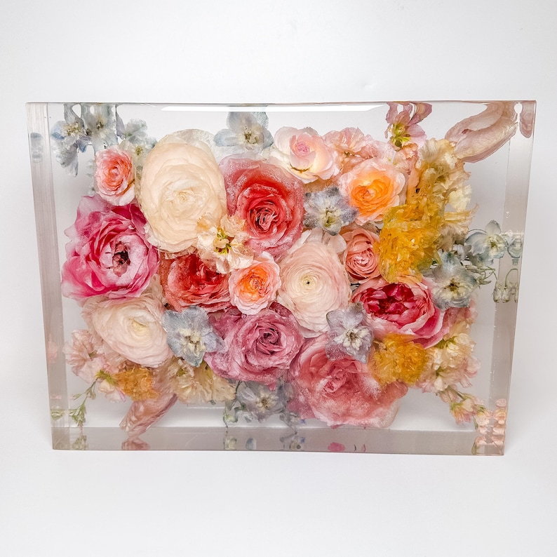 May include: A bouquet of preserved flowers in shades of pink, peach, and yellow are encased in a clear acrylic block. The flowers are arranged in a random pattern and include roses, ranunculus, and other delicate blooms.