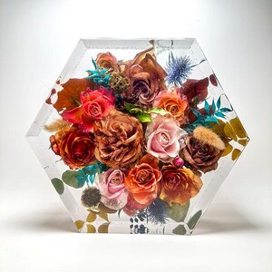 May include: A hexagonal clear resin block with a bouquet of dried flowers, including roses, in shades of red, orange, pink, and brown. The flowers are arranged in a circular pattern and are surrounded by greenery.