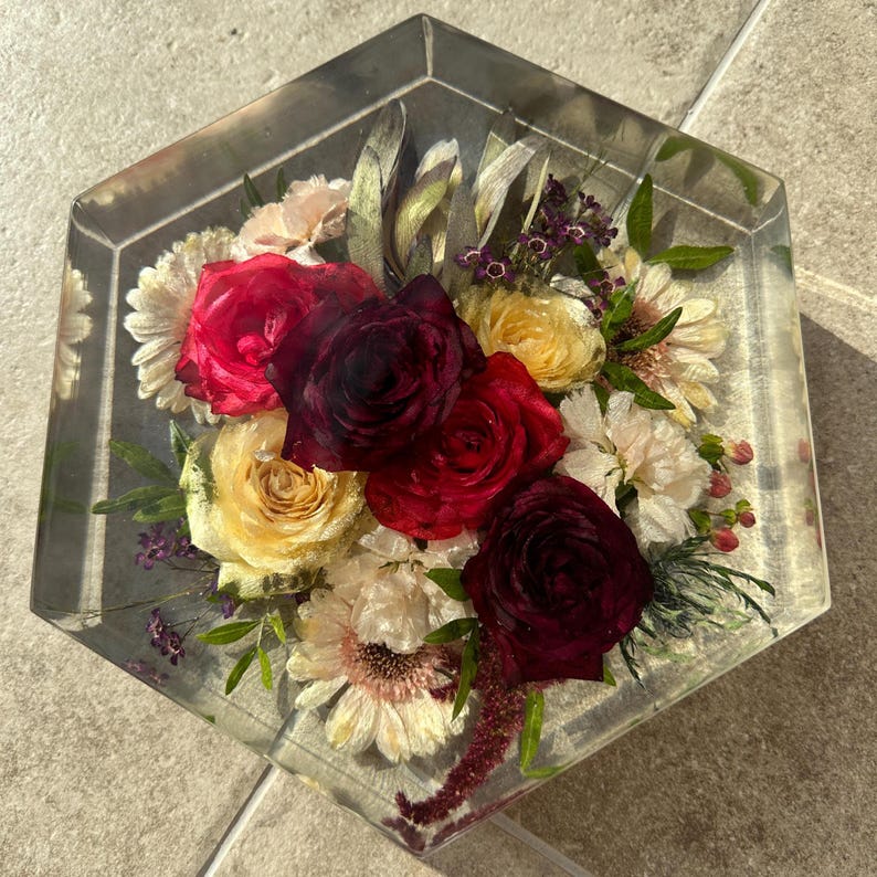 May include: A hexagonal resin block with a bouquet of preserved flowers. The bouquet includes red, burgundy, and cream roses, along with other white and purple flowers.