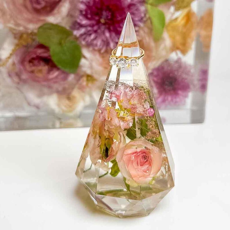 May include: A clear, faceted resin ring holder with embedded pink flowers and a gold ring with three clear stones. The ring holder is shaped like a pyramid and is filled with dried flowers. The background features a blurred floral arrangement.