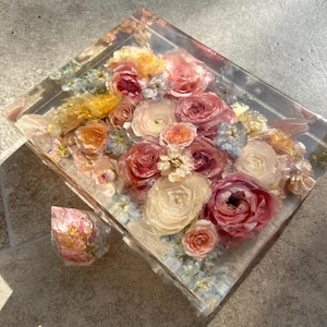 May include: A clear resin tray with a bouquet of preserved flowers. The flowers are in shades of pink, peach, and cream, with touches of yellow and blue. A smaller, matching resin piece is next to the tray.