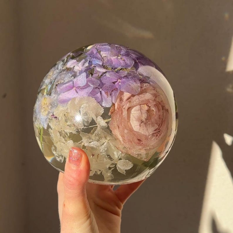 May include: A clear, spherical resin paperweight containing a variety of dried flowers. The flowers include purple hydrangeas, a pink rose, and white delicate blooms. The paperweight is held in a hand.