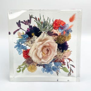 May include: A clear, square resin block encasing a vibrant floral arrangement. The bouquet features a large, pale pink rose as the focal point, surrounded by red, blue, and purple flowers, and green foliage. The flowers are preserved within the transparent block.