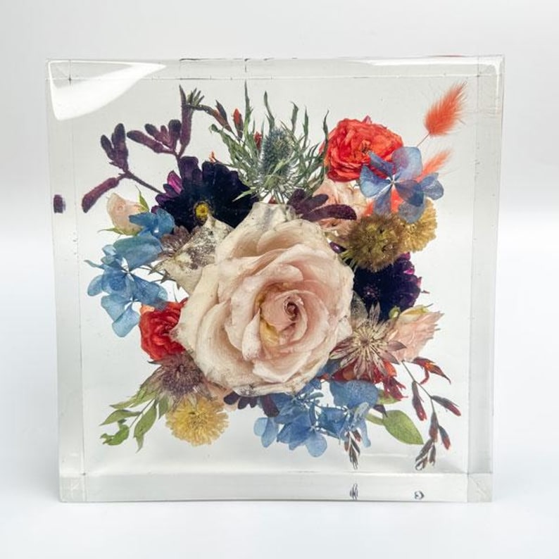 May include: A clear, square resin block encasing a vibrant floral arrangement. The bouquet features a large, pale pink rose as the focal point, surrounded by red, blue, and purple flowers, and green foliage. The flowers are preserved within the transparent block.