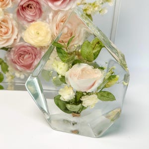 May include: A clear glass cube with a bouquet of preserved flowers inside. The flowers are a mix of white and pink roses, and green foliage.