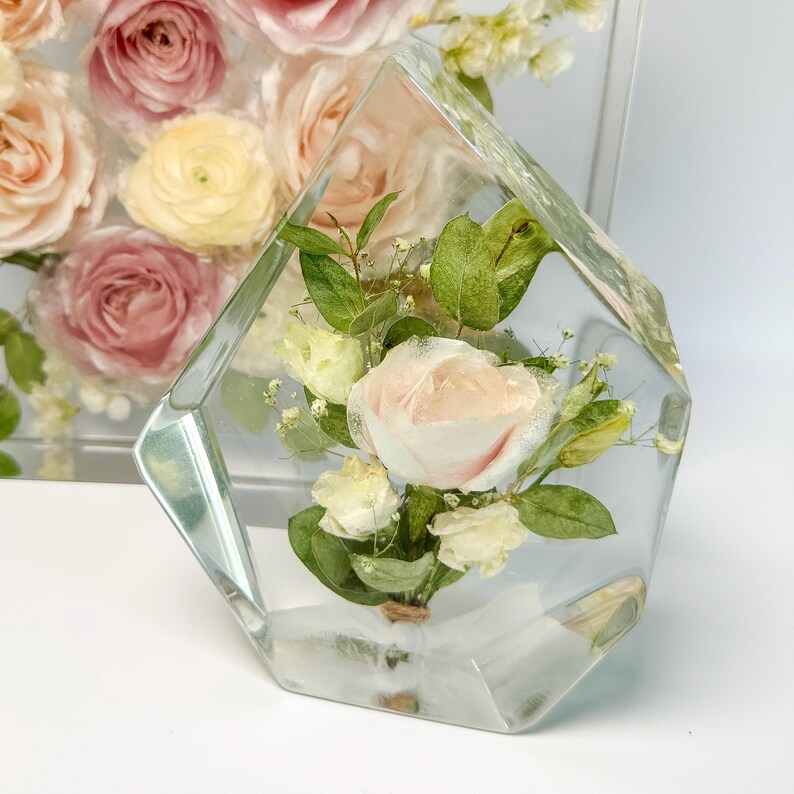 May include: A clear glass cube with a bouquet of preserved flowers inside. The flowers are a mix of white and pink roses, and green foliage.