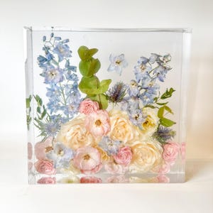 May include: A clear cube filled with preserved flowers. The flowers are a mix of light blue, pink, and white. The cube is a decorative piece for a home or office.