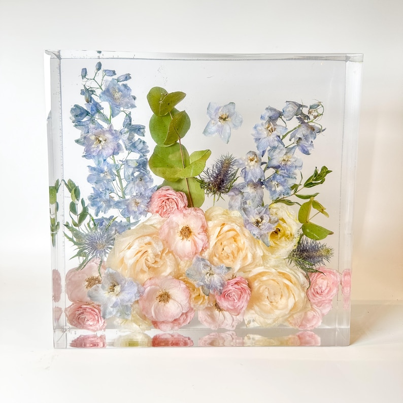 May include: A clear cube filled with preserved flowers. The flowers are a mix of light blue, pink, and white. The cube is a decorative piece for a home or office.