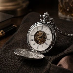 May include: A silver-toned pocket watch with a white dial featuring black Roman numerals and a skeletonised centre revealing gold-coloured mechanical gears. The watch includes a matching silver chain and a hinged cover with an intricate pattern.
