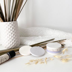 Peut inclure: Un ensemble de trois sous-verres ronds, blancs et violet clair, avec une texture naturelle et organique. Les sous-verres sont empilés les uns sur les autres et sont entourés d'herbe sèche et d'une surface blanche et moelleuse.