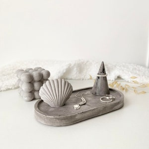 Peut inclure: Un plateau ovale en béton gris avec un cône porte-bague, une coquille et un collier et des bagues argentés. Le plateau est sur une surface blanche.