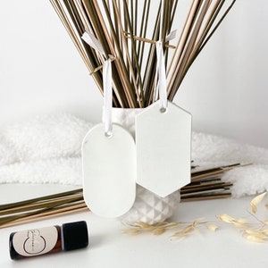 Peut inclure: Deux étiquettes décoratives blanches et vierges avec un ruban blanc sont suspendues à un vase rempli de tiges séchées. Un flacon d'huile essentielle avec un bouchon noir et une étiquette est au premier plan. Les étiquettes sont ovales et hexagonales.