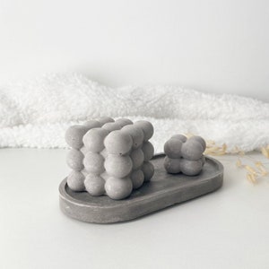 Peut inclure: Un plateau en béton gris avec deux objets en béton gris en forme de bulles. Le plus grand objet est un cube et le plus petit objet est un cube plus petit.