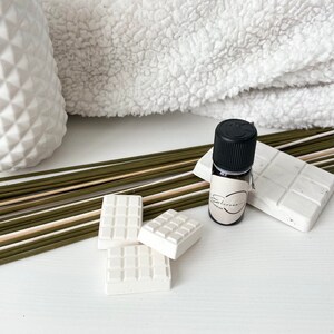 Peut inclure: Ensemble d'aromathérapie avec une petite bouteille d'huile essentielle, un bouchon noir, des barres en céramique blanches et des bâtonnets diffuseurs verts. Une serviette blanche texturée et un vase décoratif blanc sont également présents.