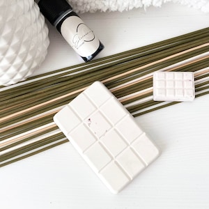 Peut inclure: Deux galets de cire blanche, l'un avec un motif en grille et l'autre avec un motif carré, sont posés sur une surface blanche avec des bâtonnets verts et bruns. Les galets de cire sont destinés à être utilisés avec des huiles essentielles.