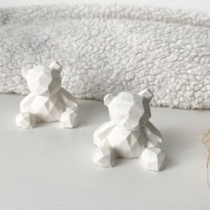 Peut inclure: Deux figurines d'ours géométriques blancs. Les ours sont assis, les jambes en avant, et présentent un design à facettes. L'arrière-plan est une surface blanche texturée.