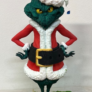 Può includere: Una statuina verde del Grinch che indossa un abito rosso da Babbo Natale con un bordo di pelliccia bianca e un cappello rosso da Babbo Natale. La statuina è in piedi su una base verde con la parola "MELODY" stampata su di essa.