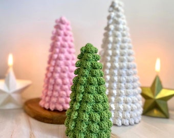 Könnte beinhalten: Drei gehäkelte Weihnachtsbäume, ein grüner, ein rosa und ein weißer, auf einer weißen Holzoberfläche. Die Bäume sind mit kleinen, runden, gehäkelten Kugeln verziert.