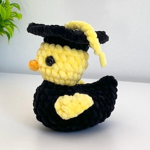 Puede incluir: Un pato amarillo tejido a ganchillo con un birrete negro con una borla amarilla. El pato tiene un ojo negro y un pico naranja. El cuerpo del pato es negro con detalles amarillos. El pato está sobre una superficie blanca.