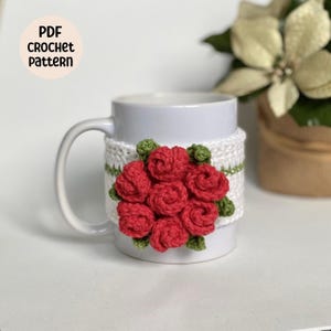 May include: White ceramic mug with a crocheted red rose flower design on the side. The mug cozy is white with green accents. The text "PDF CROCHET PATTERN" is on a pink circle in the upper left corner of the image.