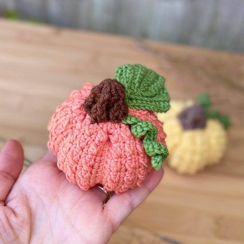 May include: A crocheted pumpkin with a green stem and brown center. The pumpkin is a light peach color.