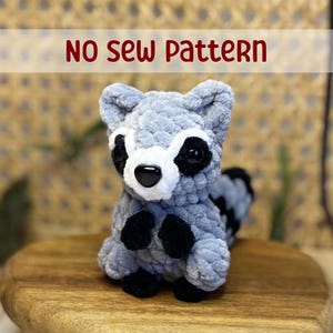 May include: A gray and black crocheted raccoon plush toy with black eyes and a white nose. The toy is sitting on a wooden surface. "NO SEW PATTERN" is written in red letters above the toy.