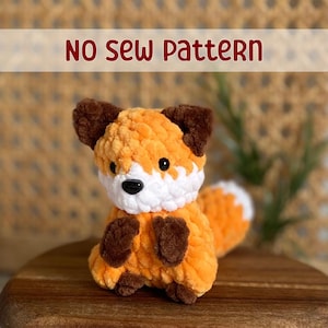May include: A crocheted fox plush toy with brown ears, a white face, and an orange body. The toy is sitting on a wooden surface. The text "NO SEW PATTERN" is displayed above the toy.