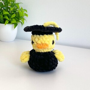 May include: A handmade, yellow and black crocheted duck wearing a black graduation cap with a yellow tassel. The duck has a yellow body, black eyes, and an orange beak. The plushie is on a white surface with a small green plant in the background.