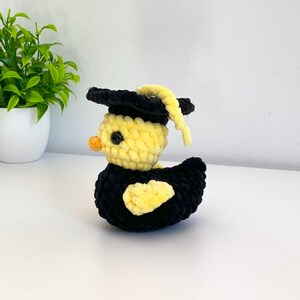 May include: A crocheted yellow duck wearing a black graduation cap with a yellow tassel. The duck has a black eye and an orange beak. The duck's body is black with yellow accents. The duck is sitting on a white surface.