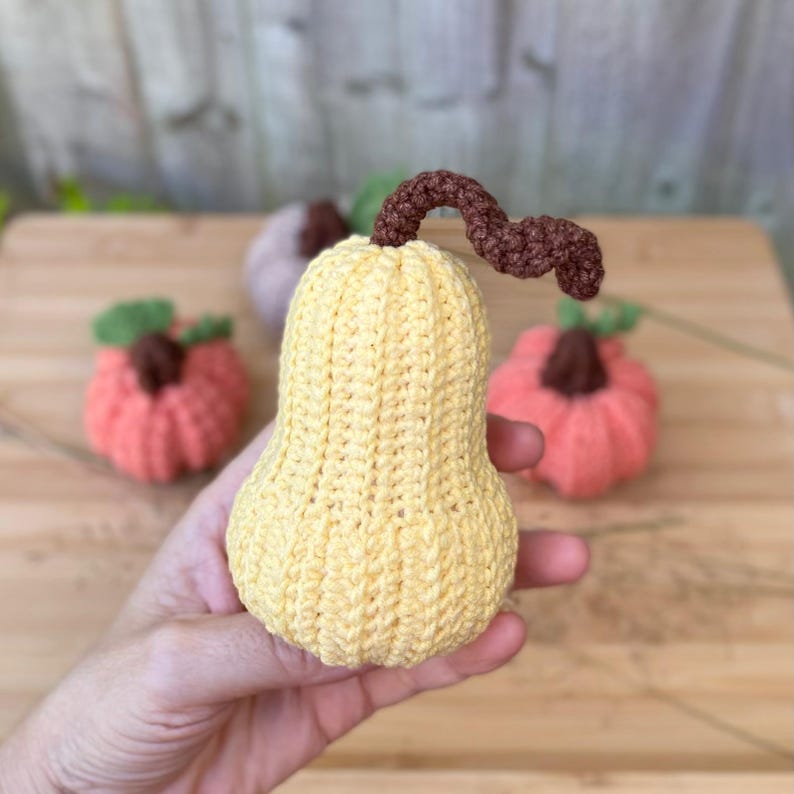 May include: A crocheted yellow gourd with a brown stem. The gourd is sitting on a wooden surface with two other crocheted gourds, one pink and one purple.