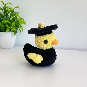May include: A handmade, plush duck wearing a black graduation cap. The duck has a yellow body, black wings, and an orange beak. The duck is sitting on a white surface.