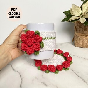 May include: White crocheted mug cozy with red roses and green leaves. The cozy is on a white mug. A white crocheted coaster with red roses and green leaves is on a marble surface. The text "PDF CROCHET PATTERN" is on a pink circle.
