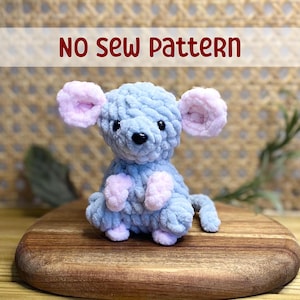May include: A no-sew pattern for a crocheted mouse toy. The mouse is light blue with pink ears, paws, and tail. The mouse is sitting on a wooden surface.