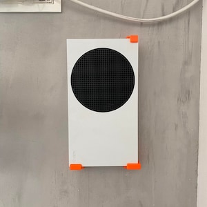 May include: A white Xbox Series X console mounted on a wall with orange brackets. The console has a black circular vent on the front.