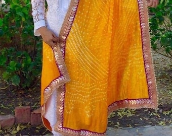 May include: A yellow dupatta with a white and gold embroidered border. The dupatta has a paisley pattern and is made of a sheer fabric.