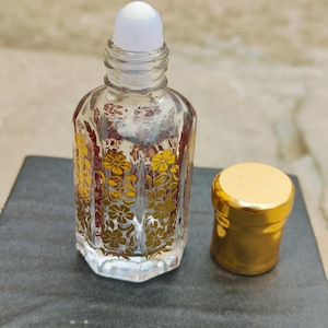 May include: A clear glass perfume bottle with a white rollerball top and a separate gold-colored cap. The bottle is decorated with a gold floral pattern. The bottle is sitting on a dark surface.