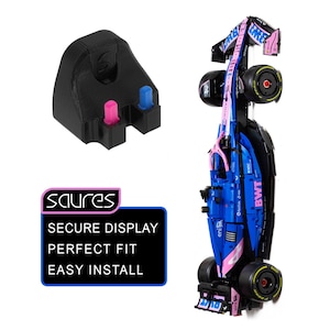May include: A black plastic wall mount bracket with two small pink and blue pegs, shown alongside a blue and pink Formula 1 race car model. A black rectangular sign features the white text: saures, SECURE DISPLAY, PERFECT FIT, EASY INSTALL.