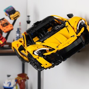 May include: A yellow and black Lego model car, a McLaren Senna, is displayed on a white wall. The car is suspended in mid-air, as if it is flying.