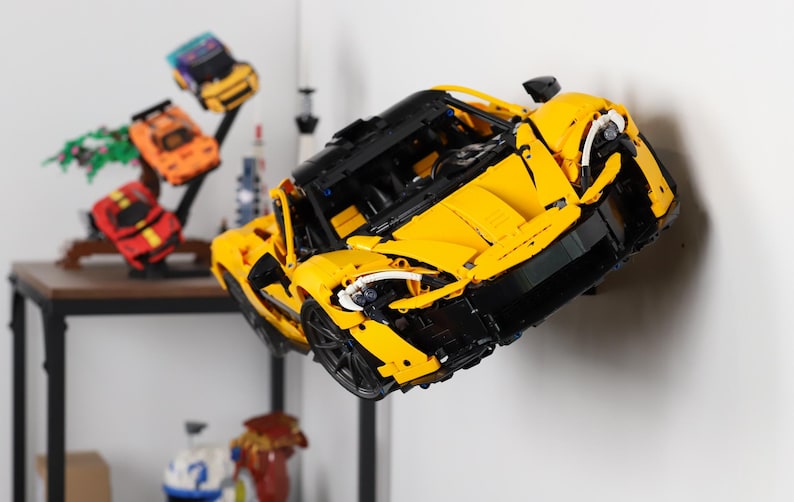May include: A yellow and black Lego model car, a McLaren Senna, is displayed on a white wall. The car is suspended in mid-air, as if it is flying.