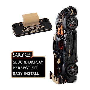 May include: A black and tan model sports car positioned vertically next to a black rectangular display plaque with the text KOENIGSEGG SADAIR'S SPEAR 42232. Below, a black sign with orange trim displays the text SAURES SECURE DISPLAY PERFECT FIT EASY INSTALL.