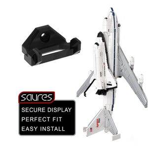 May include: A black 3D-printed display mount next to a white and gray model of a NASA space shuttle. The shuttle has the NASA logo and USA markings. The mount is designed to hold the shuttle. The image also includes a graphic with the text "SECURE DISPLAY PERFECT FIT EASY INSTALL".