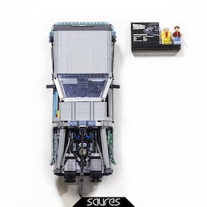 Puede incluir: Un DeLorean de Lego gris y azul de la película Regreso al futuro. El coche está construido con ladrillos Lego y tiene un interior detallado. El coche está sentado sobre una superficie blanca. La caja del set de Lego está en el fondo. La caja es negra con texto blanco y una imagen del DeLorean.