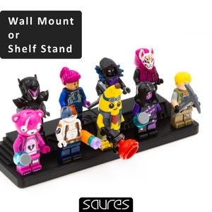 Peut inclure: Un présentoir noir avec huit figurines, dont une figurine rose et blanche, une figurine jaune, une figurine violette, une figurine noire, une figurine bleue, une figurine rouge, une figurine verte et une figurine jaune. Le présentoir porte l'inscription "Wall Mount or Shelf Stand" sur le devant.