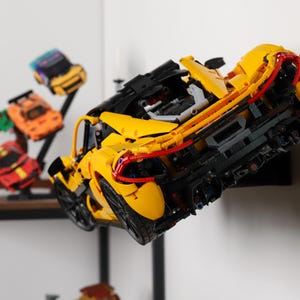 May include: A yellow, red, and black Lego McLaren Senna sports car model is mounted on a black wall mount. The car is displayed with other smaller Lego car models on a shelf.