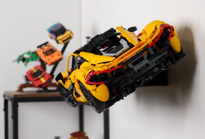 May include: A yellow, red, and black Lego McLaren Senna sports car model is mounted on a black wall mount. The car is displayed with other smaller Lego car models on a shelf.